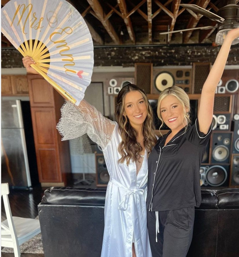 A bride in a satin robe and her friend holding up a custom white and gold Daftboy Mrs. wedding fan