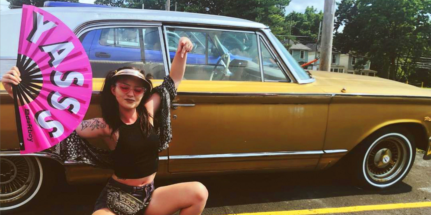 A customer holding a pink Daftboy Yasss hand fan in front of a vintage gold car