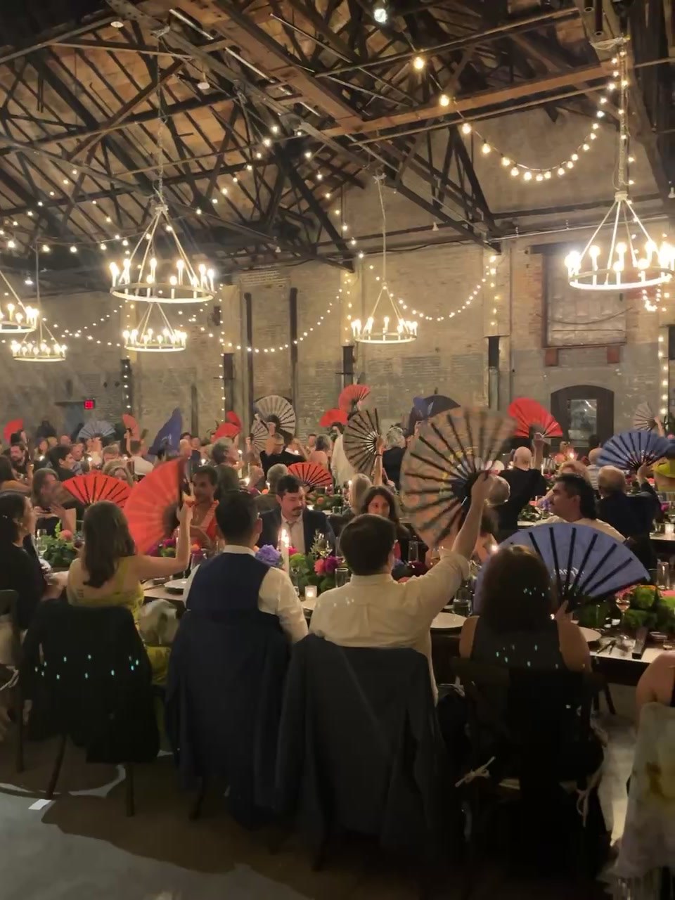 A candlelit reception hall full of wedding guests waving custom Daftboy paisley fans